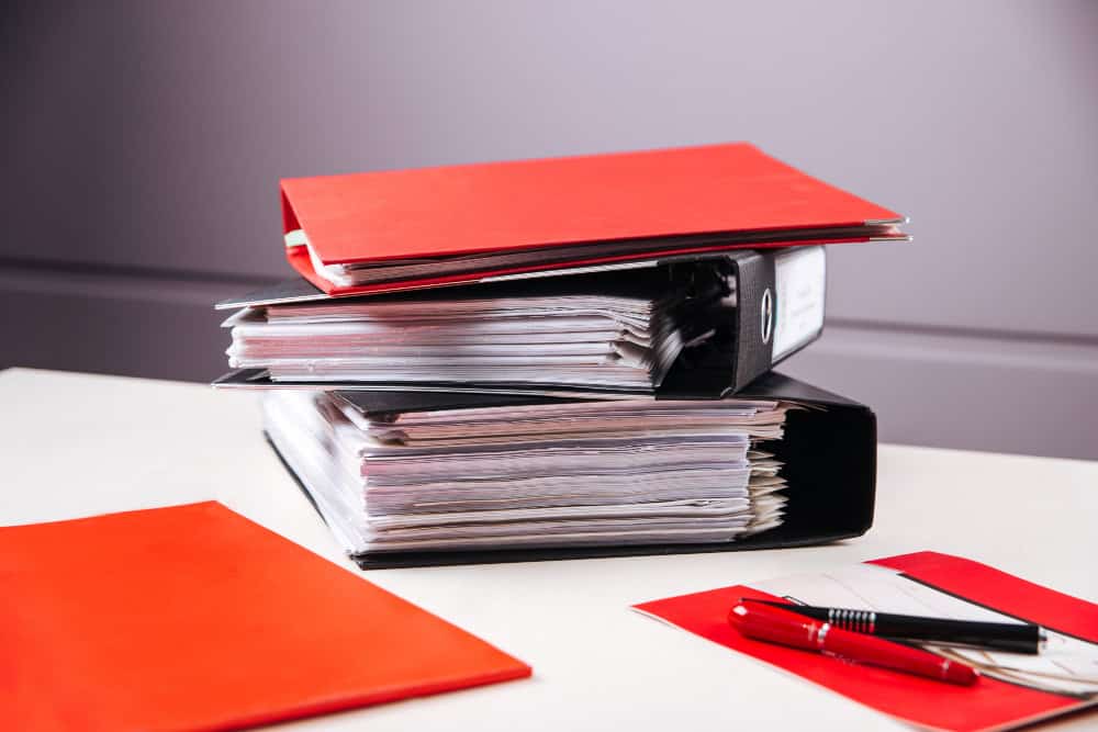 Trois classeurs, deux noirs et un rouge, remplis de documents papier sont empilés sur un bureau blanc. Un dossier rouge, un stylo et d'autres papiers se trouvent également sur le bureau. L'arrière-plan est uni et flou.