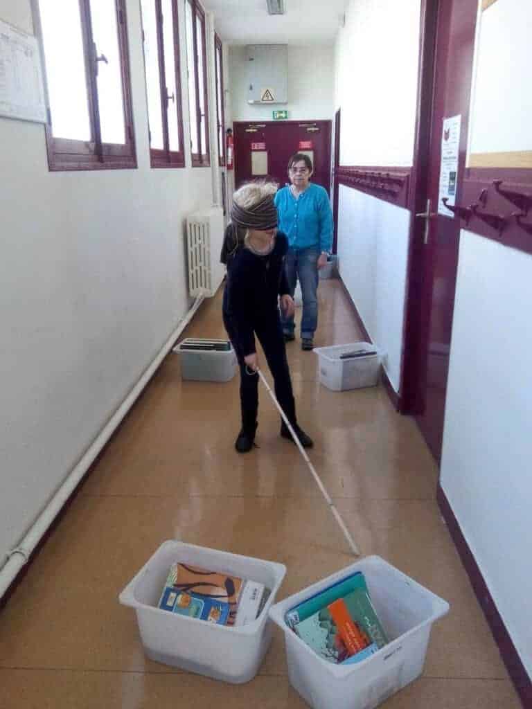 Un enfant portant un masque pour les yeux utilise une canne blanche pour se déplacer dans un couloir, se déplaçant entre des bacs en plastique contenant des livres, tandis qu'un adulte observe par derrière.
