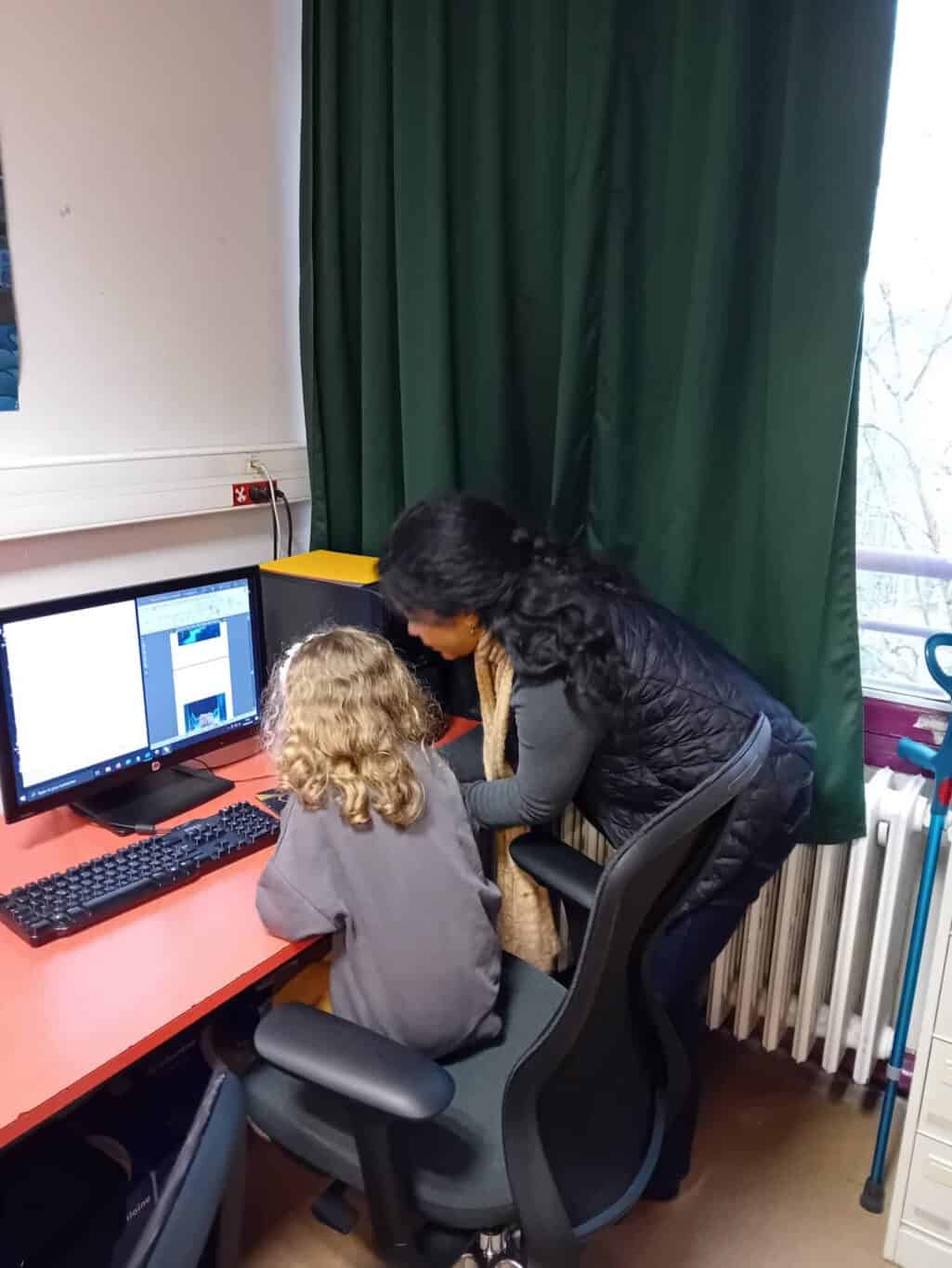 Une femme se penche pour aider un enfant aux cheveux longs et bouclés qui est assis à un bureau d'ordinateur dans une salle de classe, tous deux regardant l'écran de l'ordinateur.
