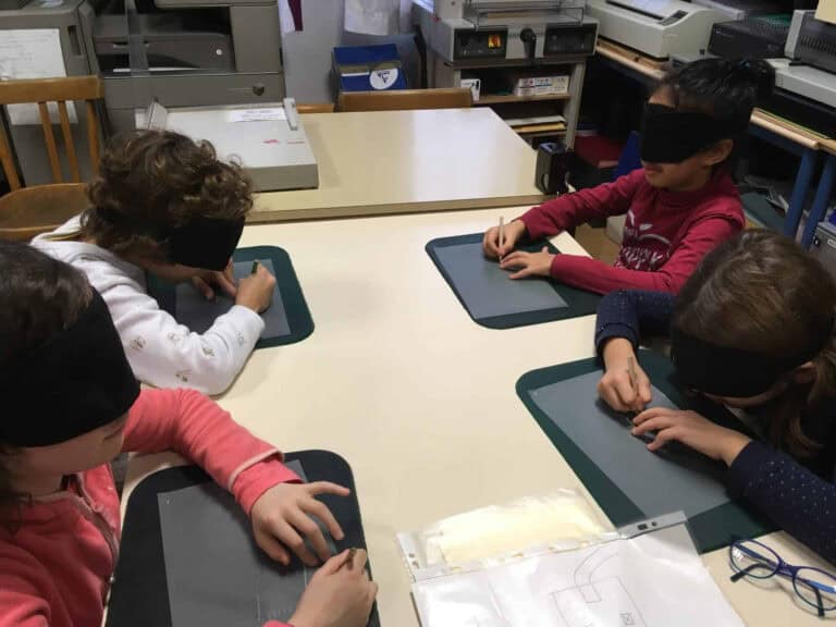 Quatre enfants portant des bandeaux sur les yeux sont assis autour d'une table, utilisant leurs mains pour sentir et tracer sur des tableaux tactiles, participant éventuellement à une activité d'apprentissage tactile ou en Braille.