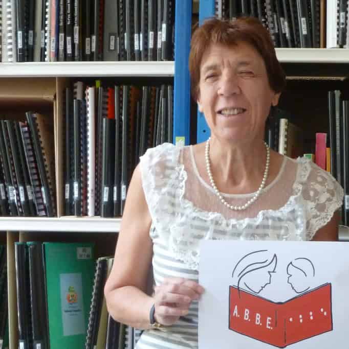 Une femme aux cheveux bruns courts et portant un haut en dentelle blanche sourit tout en tenant une pancarte avec une illustration de deux visages au-dessus d'un livre intitulé « A.B.B.E. » devant des étagères remplies de livres et de dossiers.