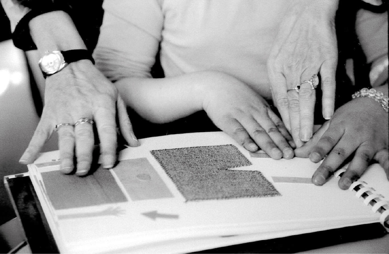 Trois mains – deux adultes et un enfant – touchent ensemble les pages texturées d'un livre, se livrant peut-être à une lecture tactile. L'image, en noir et blanc, présente des détails rapprochés des mains et des bagues.
