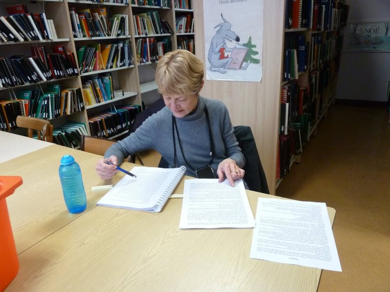 Une femme est assise à une table dans une bibliothèque, lisant et corrigeant des devoirs. Des étagères remplies de livres se trouvent à l'arrière-plan. Une bouteille d'eau bleue et d'autres papiers sont posés sur la table.