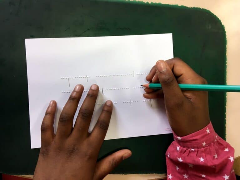 Une personne portant une chemise rose à motifs d'étoiles utilise un crayon vert pour suivre des points braille en relief sur une feuille de papier blanc posée sur une surface vert foncé.
