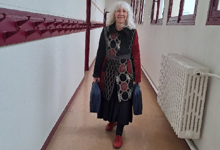 Une femme âgée aux longs cheveux gris, portant un manteau coloré à motifs et des chaussures rouges, sourit en marchant dans un couloir avec des crochets et des fenêtres marron, portant deux sacs sombres.